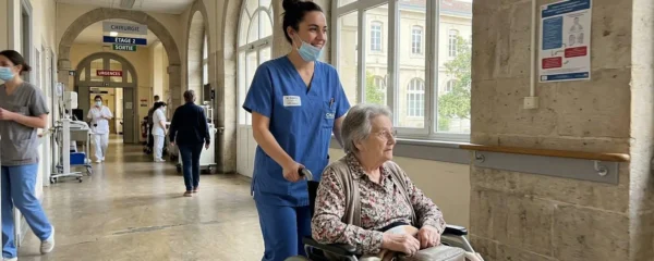 Une infirmière souriante accompagne une résidente âgée en fauteuil roulant dans un couloir lumineux avec architecture moderne.