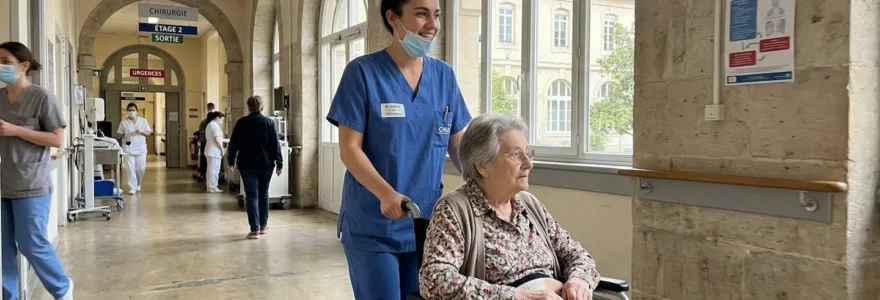 Une infirmière souriante accompagne une résidente âgée en fauteuil roulant dans un couloir lumineux avec architecture moderne.